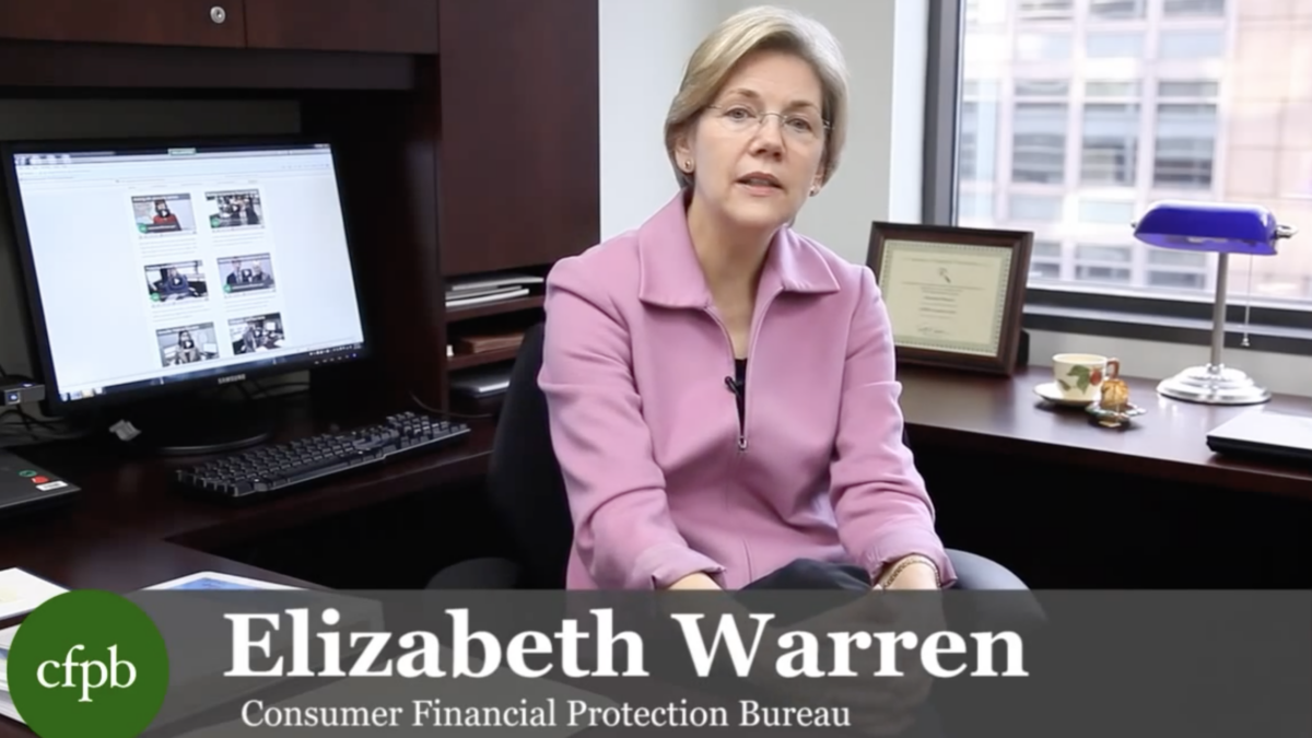 Elizabeth Warren sits behind desk in pink jacket