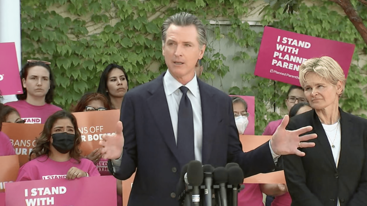Gavin Newsom speaking in front of crowd of angry women holding planned parenthood signs