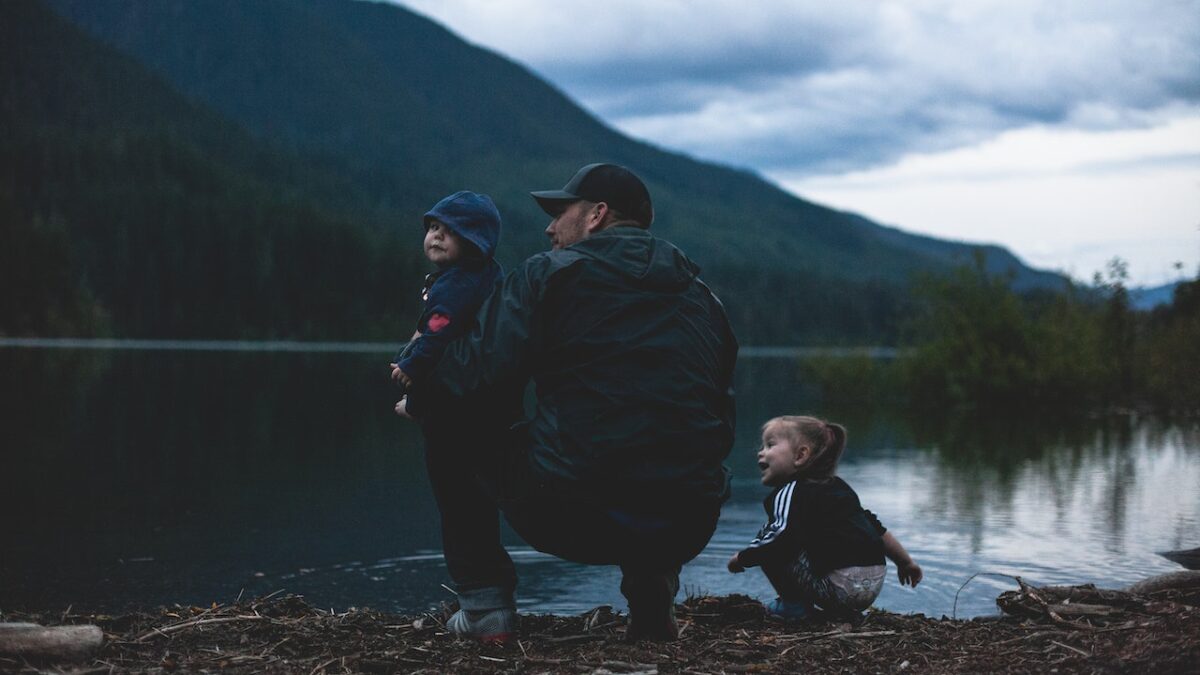 man with two children crouching beside a lake in the mountains