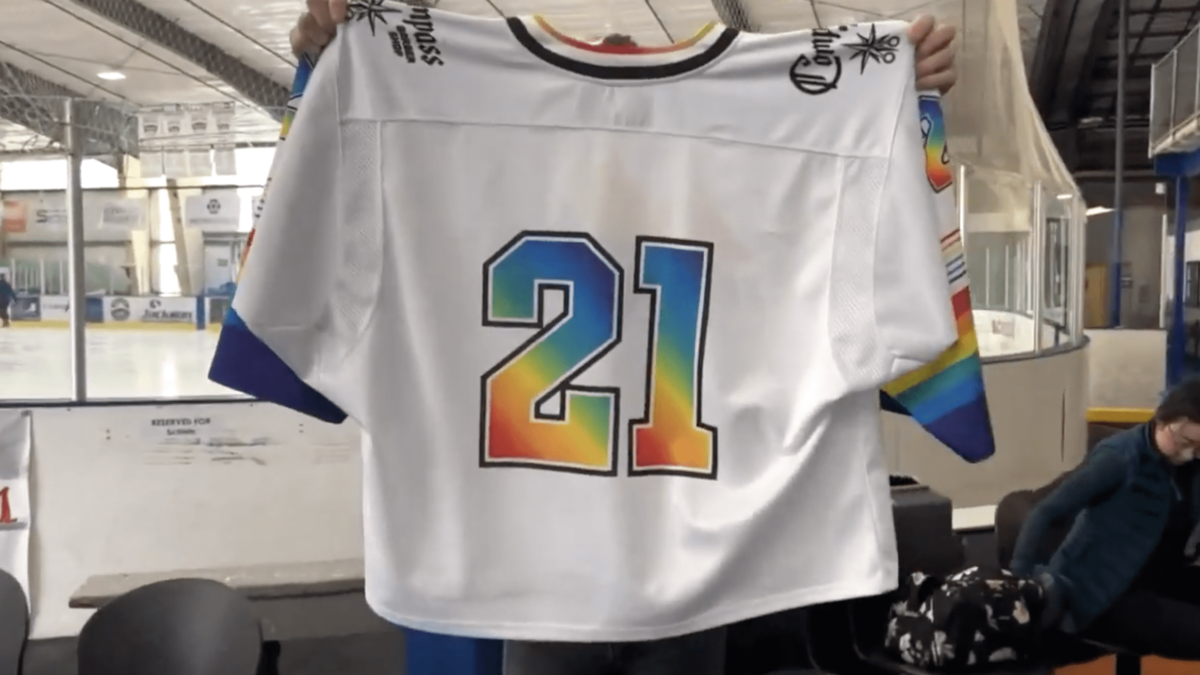 man holding up rainbow "pride" themed hockey jersey inside hockey rink