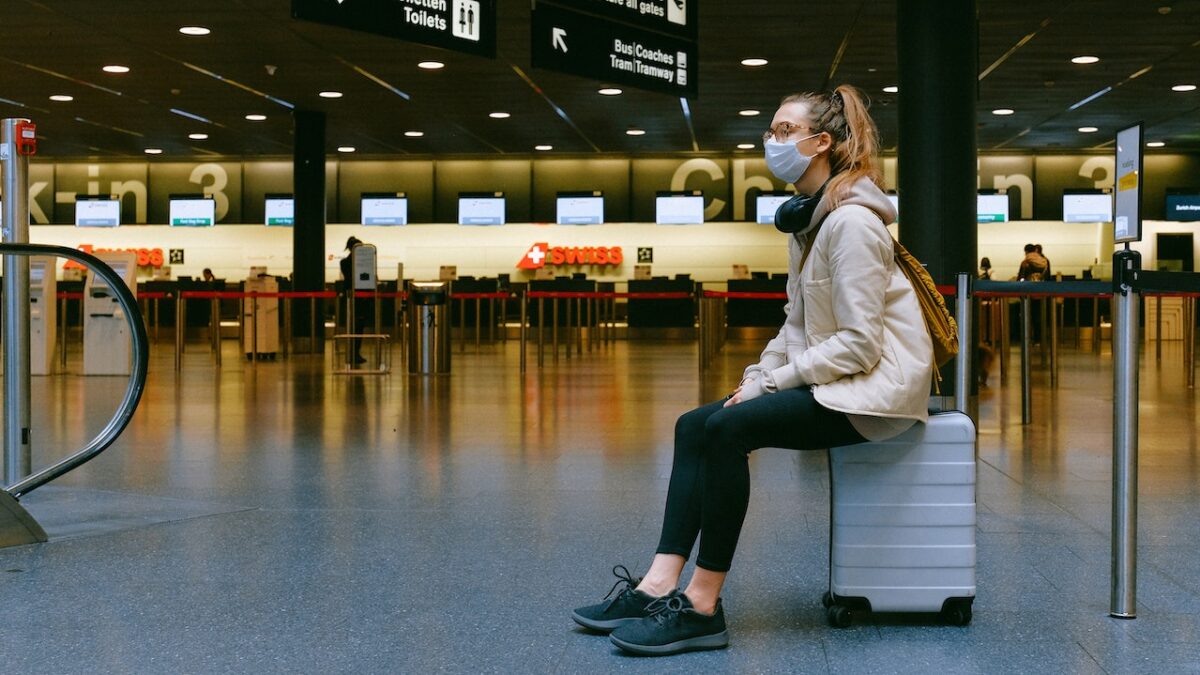 woman wearing a mask in an airport and sitting on her suitcase