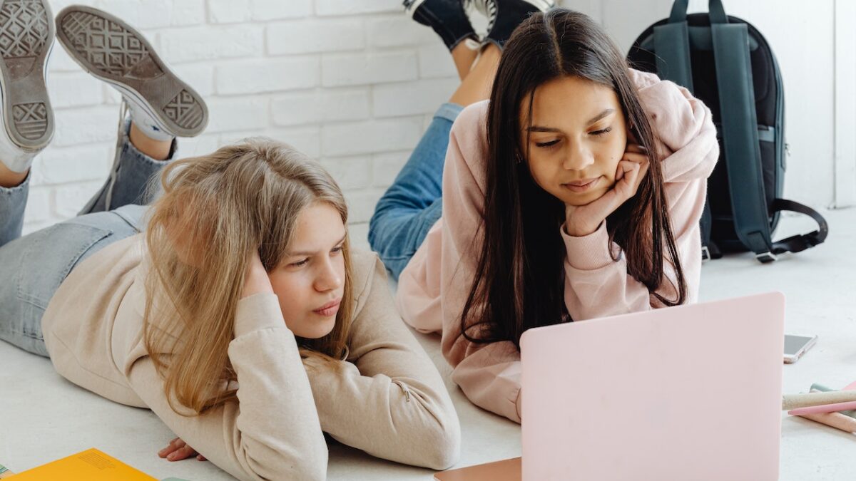 two high school girls lying on the ground looking at a laptop