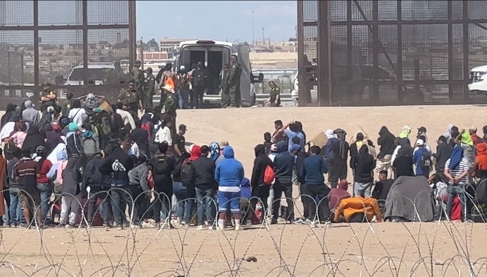 group of migrants gathered at border point of entry behind wire fence