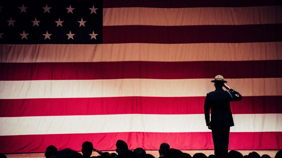 Service member facing and saluting the American Flag, other service members behind him.