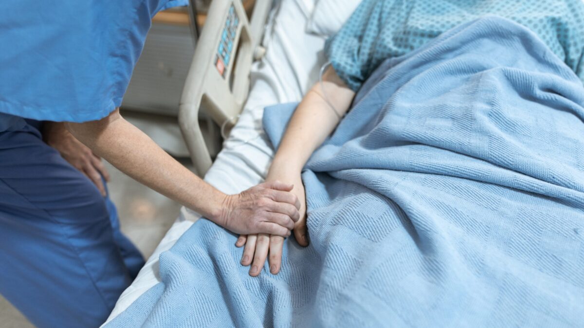 Patient in hospital bed while someone bedside holds their hand.