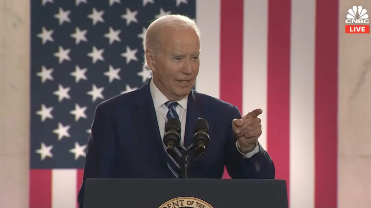 Joe Biden behind a podium pointing at the audience, American flag behind him.