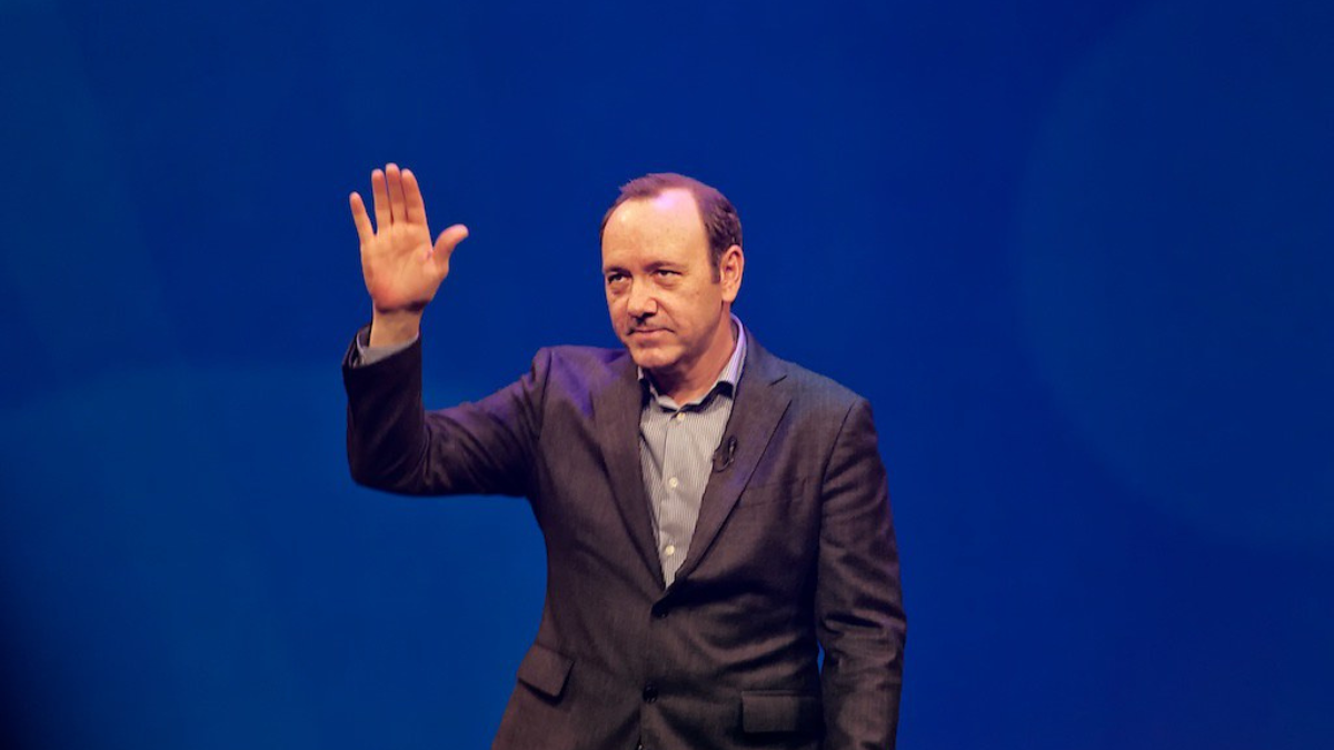 Kevin Spacey standing in front of a blue background waving
