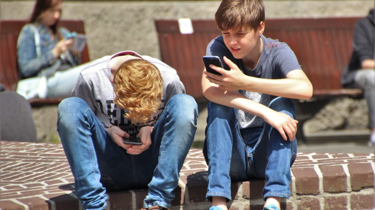 Two boys in the foreground sitting on a step looking at their phones. One girl in the background on her phone