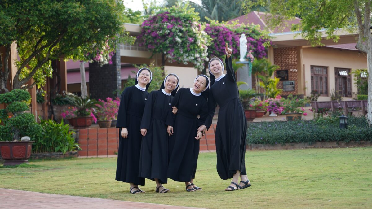 Four nuns stand together in a courtyard. One points upward while the other look up.