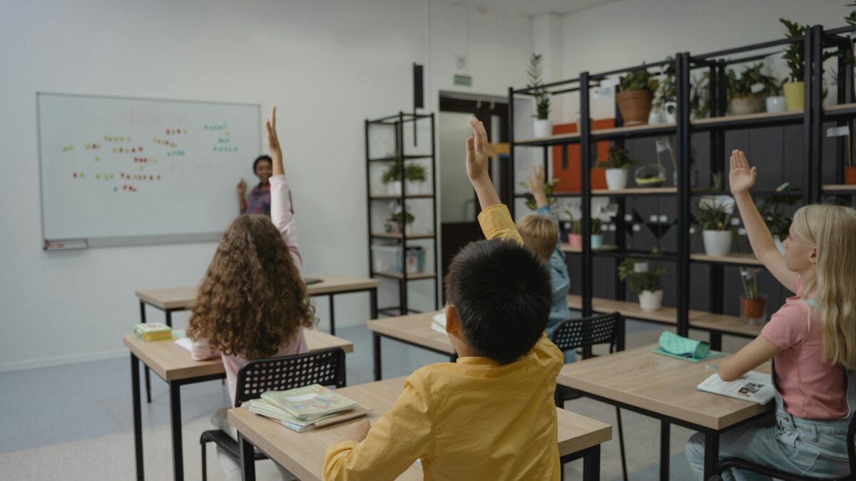 Four students in a classroom raise their hands while teacher is at the front of the room.