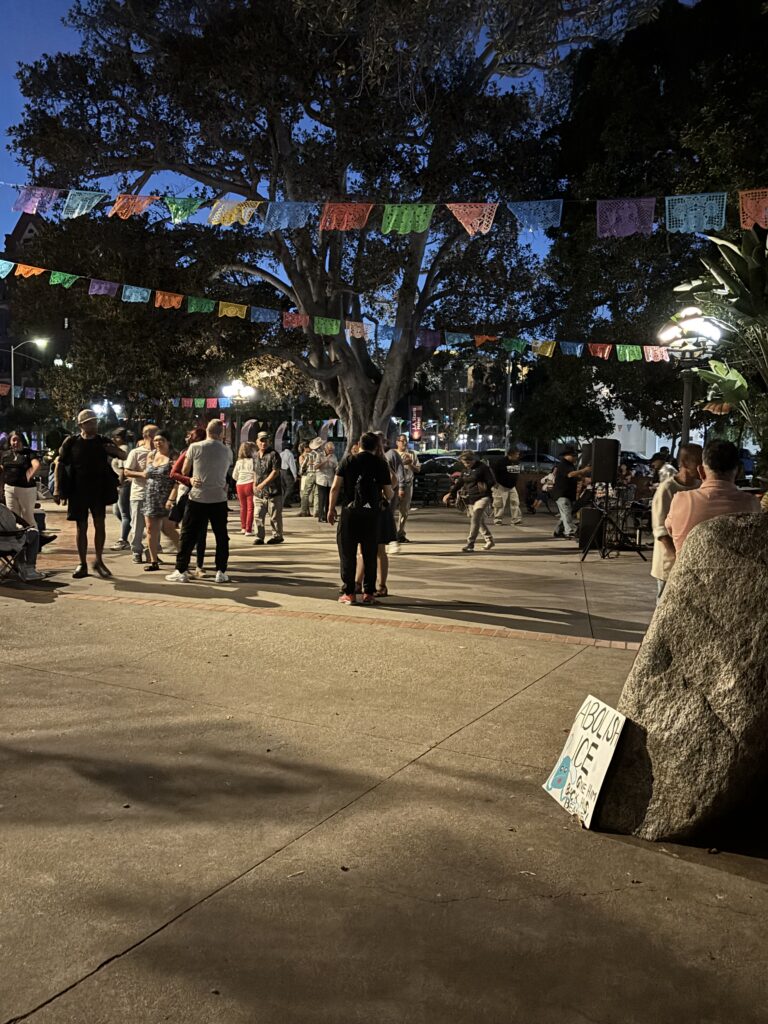 A small and peaceful crowd dances beneath brightly colored flags during a pleasant evening.