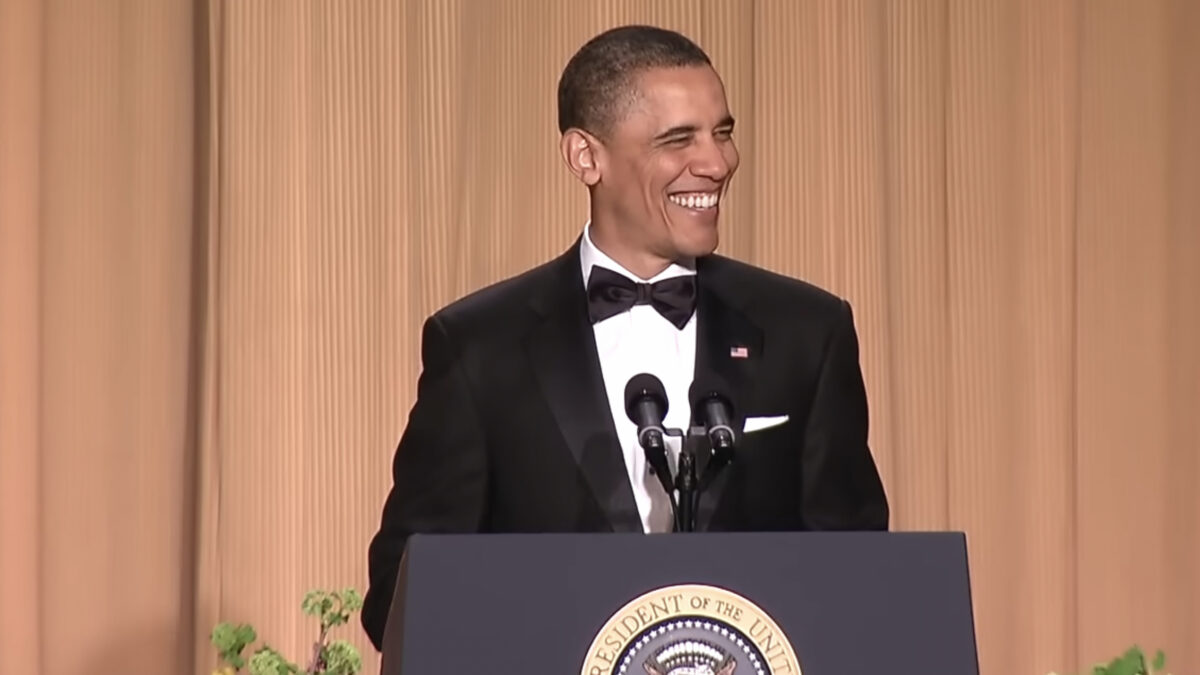 Barack Obama, grinning broadly, wears a tuxedo int front of a plain brown backdrop, behind a lectern with the seal of the President of the Uinted States.