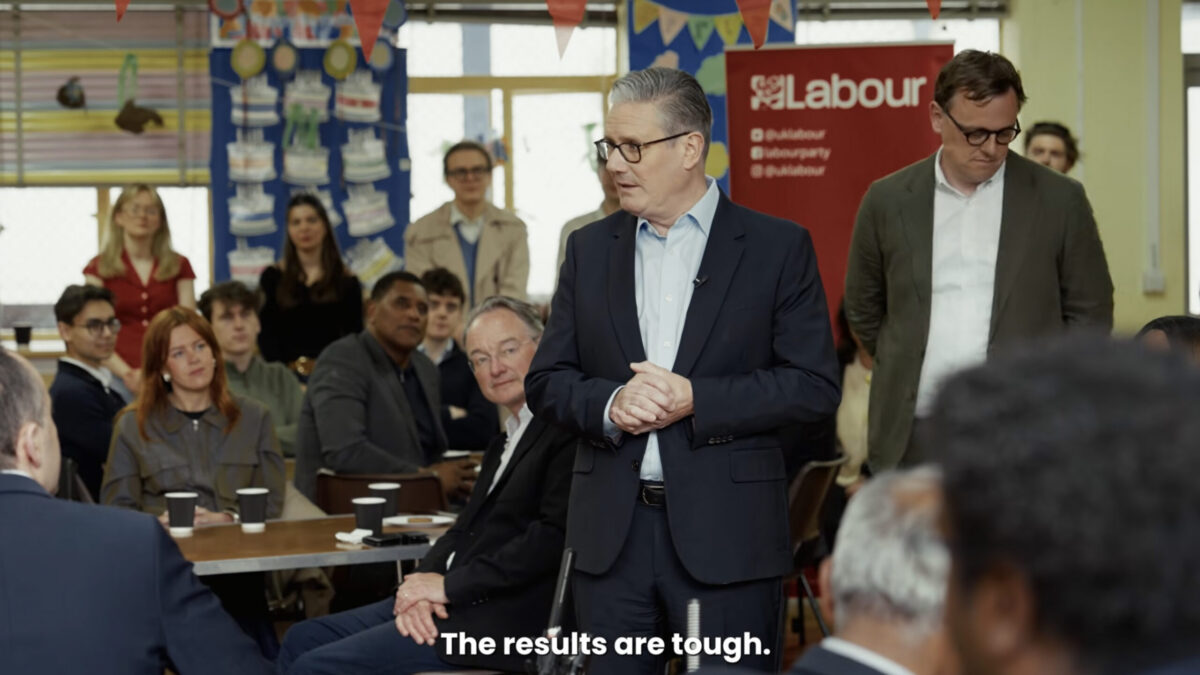 In a crowded room, in front oid a red Labour Party banner, British Prime Minster Keir Starmer rubs his hands together and looks worried as he speaks.