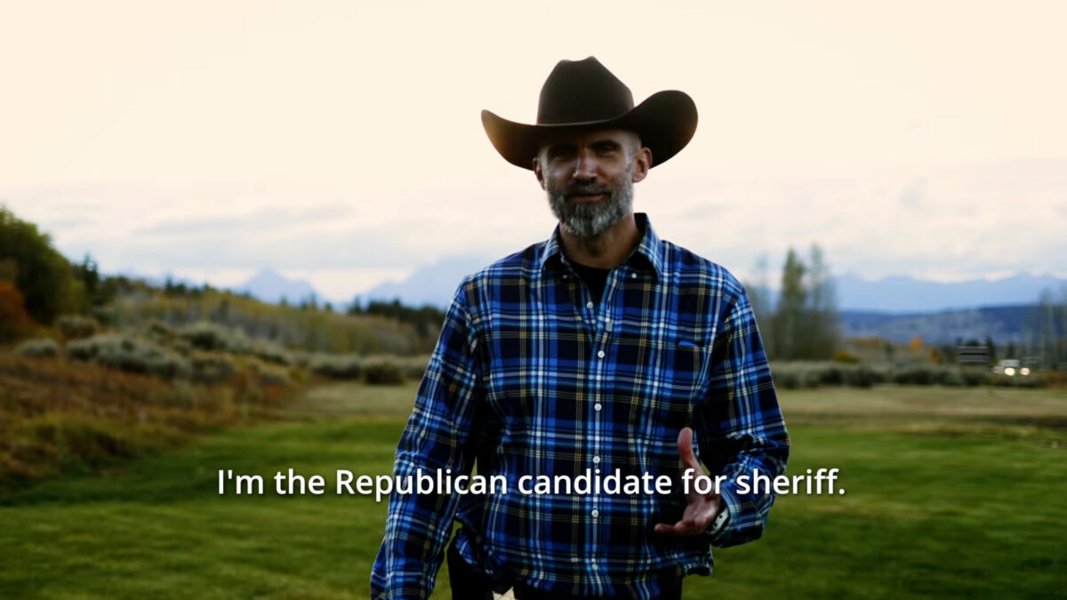 A bearded man in a flannel shirt and cowboy hat walks in a grassy field, over a caption that identifies him as the Republican candidate for sheriff.
